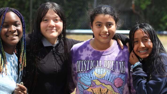 Four students standing together outside, arms over each other shoulders, all smiling at camera